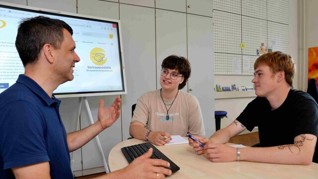 Ein Mitarbeiter der Pädagogischen Hochschule Heidelberg sitzt mit zwei Studierenden am Tisch. Der Mitarbeiter spricht deutlich mit den Händen. Die Studentin und der Student hören ihm aufmerksam zu. Das Bild wurde in der Vertrauensstelle für studentische Beschwerden aufgenommen.