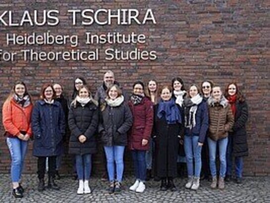 Studierende und Dozierende stehen vor einer Backsteinwand. Darauf steht geschrieben: Klaus Tschira - Heidelberg Institute for Theoretical Studies