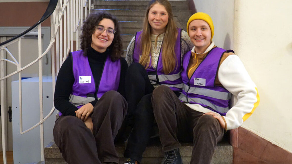 Das studentische Awarness-Team sitzt auf einer Treppe und lacht in die Kamera. Man erkennt sie an den lila Westen.