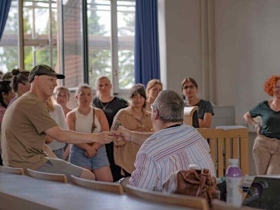 Das Bild zeigt eine Gruppe Studierender, die mit Bildungsfachkraft Helmuth Pflantzer im Gespräch sind. Ein Student reicht Herrn Pflantzer, der von hinten im Bild ist, ein Mikrofon.