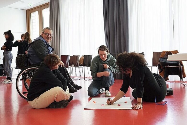 Mehrere Menschen sitzen auf dem Boden oder im Rollstuhl um ein Flip-Chart-Papier herum. Eine Person schreibt etwas auf das Papier.