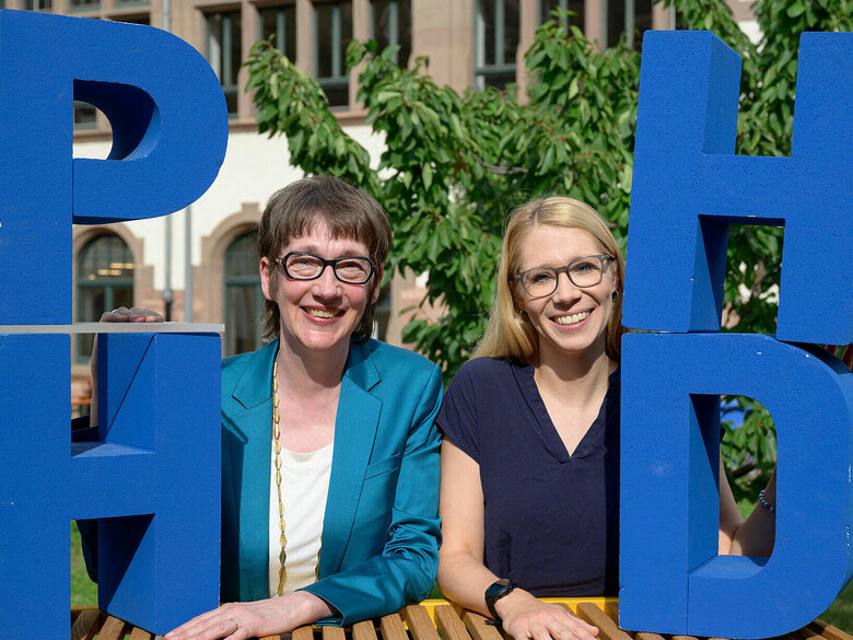 Karin Vach und Tanja Eschenauer-Engler im Innenhof der Pädagogischen Hochschule Heidelberg. Sie stehen zwischen Styroporbuchstaben "PHHD".