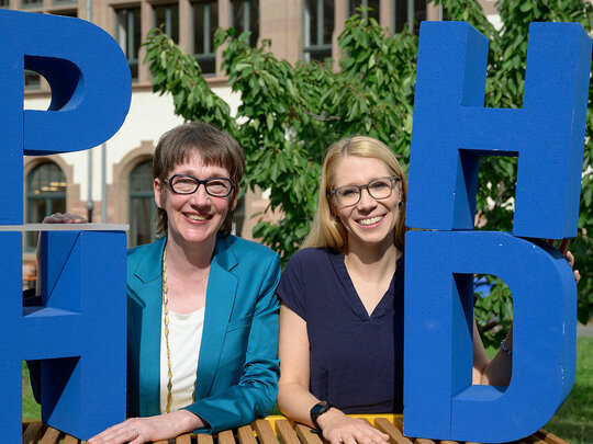 Karin Vach und Tanja Eschenauer-Engler im Innenhof der Pädagogischen Hochschule Heidelberg. Sie stehen zwischen Styroporbuchstaben "PHHD".