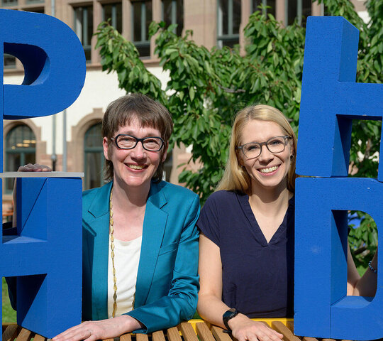 Karin Vach und Tanja Eschenauer-Engler im Innenhof der Pädagogischen Hochschule Heidelberg. Sie stehen zwischen Styroporbuchstaben "PHHD".