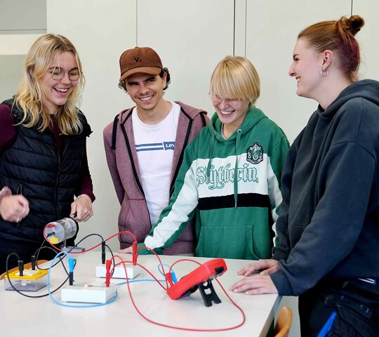 Drei Studentinnen und ein Student stehen um einen Tisch auf dem ein physikalischer Versuch aufgebaut ist.