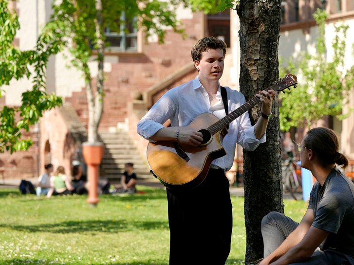 Zwei Studierende sitzen vor dem Altbau und musizieren zusammen. 