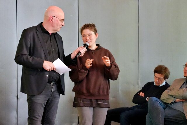 Zu sehen ist Moderator Armin Himmelrath sowie jemand aus dem Publikum, der/die eine Frage an das Podium stellt.