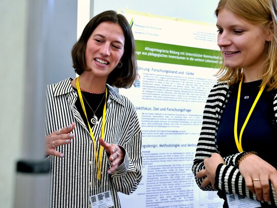 Das Foto zeigt zwei Personen, die sich vor einem Plakat unterhalten.