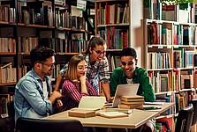 Das Bild zeigt eine Gruppe junger Menschen gemeinsam an einem Tisch. Im Hintergrund befinden sich mit Büchern gefüllte Regale.